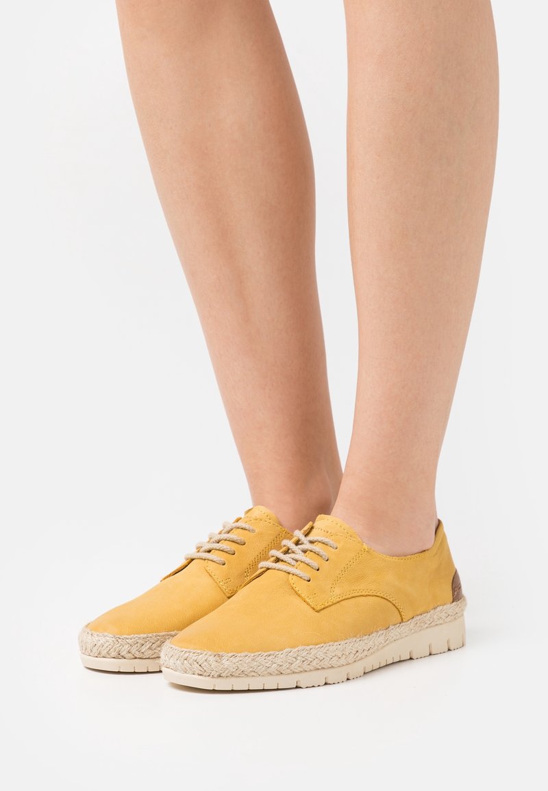Personne portant des chaussures décontractées jaunes à lacets avec garniture en corde tressée et semelles blanches, visible de mi-mollet vers le bas sur un fond blanc.