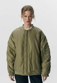Olivenfarvet bomberjakke med en quiltet front, ribbet krave og elastiske ærmer. Har en sidelomme med lynlås og et afslappet fit design.