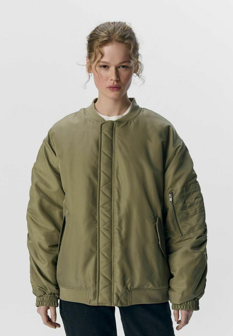 Olivenfarvet bomberjakke med en quiltet front, ribbet krave og elastiske ærmer. Har en sidelomme med lynlås og et afslappet fit design.