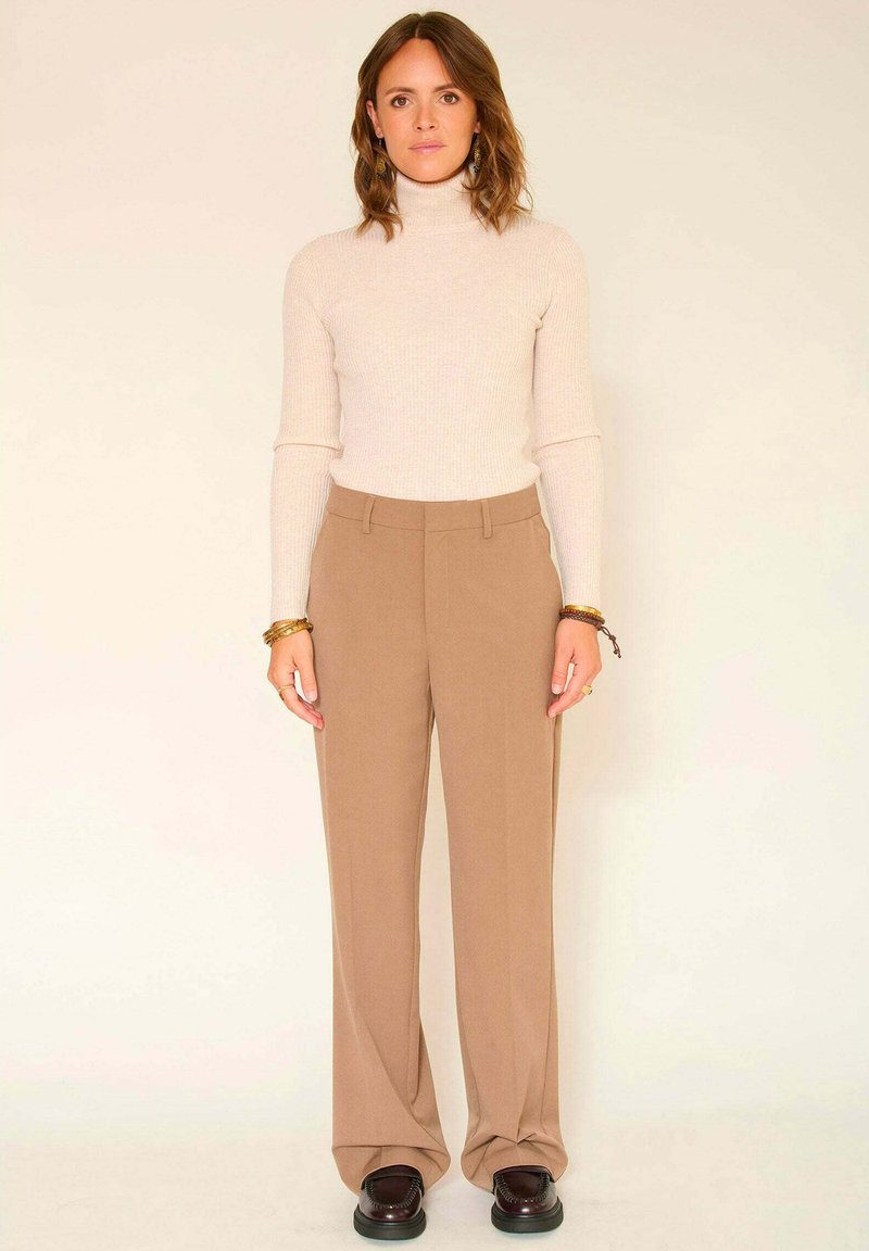 Un pull col roulé en maille côtelée crème associé à un pantalon large taille haute beige, doté d'une texture lisse et d'une coupe ajustée, complété par des chaussures foncées.