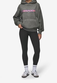 Grå oversized hoodie med "PEGADOR" tryckt i livfulla färger, tillsammans med svarta leggings. Modellen bär även vita strumpor och grå sneakers.