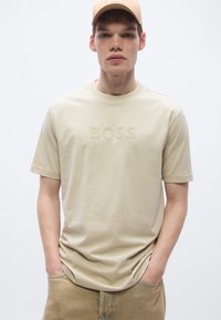 Bézs pamut póló rövid ujjakkal és kerek nyakkivágással; enyhe, dombornyomott "BOSS" logóval az elülső részen.
