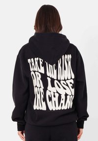 Zwarte hoodie met witte tekst op de achterkant die luidt: "NEEM HET RISICO OF VERLOOR DE KANS." Zacht materiaal, capuchon met trekkoord en geribbelde manchetten.