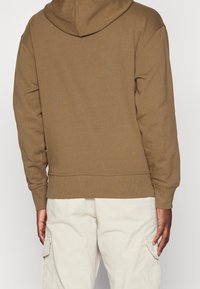 Felpa con cappuccio marrone con finitura testurizzata, cappuccio regolabile, polsini e orlo a coste. Indossata con pantaloni cargo di colore chiaro. Vista posteriore semplice.