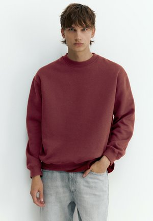Junger Mann mit braunen Haaren, der einen dunkelroten Sweatshirt und hellgraue Jeans trägt, steht vor einem schlichten weißen Hintergrund mit einer Hand in der Hosentasche.