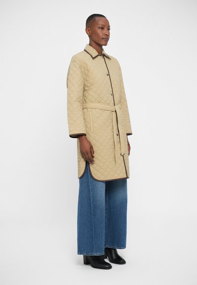 Quilted beige coat with a belted waist, brown trim, button front, and side slits. Paired with wide-leg blue denim jeans and black ankle boots.