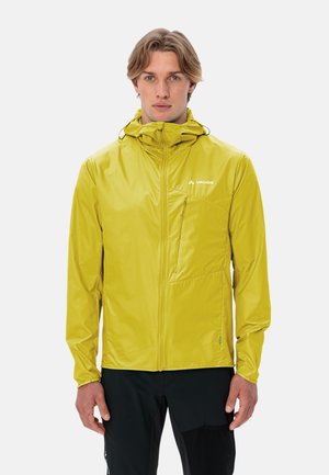 Junger Mann, der nach vorne blickt und eine limettengrüne, mit Reißverschluss versehene Jacke mit Kapuze sowie schwarze Hosen trägt, vor einem schlichten weißen Hintergrund.