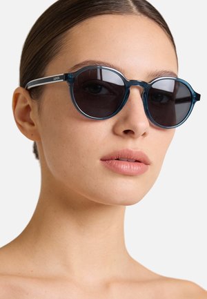 Occhiali da sole rotondi con lenti scure e montatura trasparente blu. Accenti argentati sulle astine. Design pulito con dettagli minimalisti.