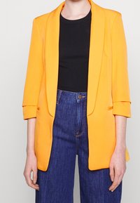 Oranžový blazer s přizpůsobeným designem, strukturovanými klopami a rukávy po tři čtvrtiny, spárovaný s černým topem a džínovými kalhotami s vysokým pasem.