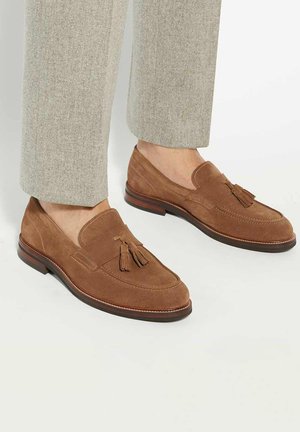 Brown suede loafers with tassel detail, rounded shape, and a low stacked heel. Featured with contrasting stitching on the upper.