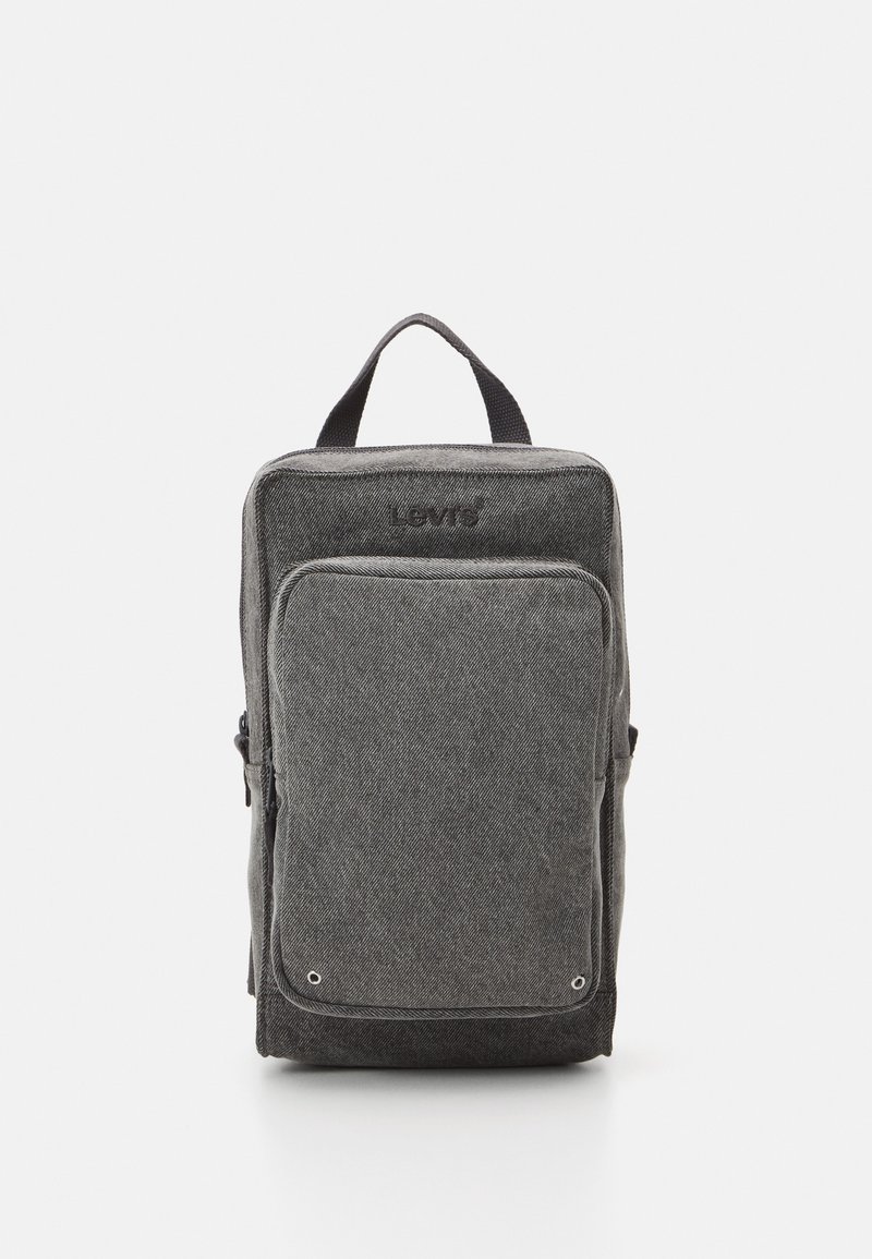 Mochila de tela gris con forma rectangular, bolsillo frontal con cremallera y detalle de logo. Presenta un asa superior y una superficie texturizada.
