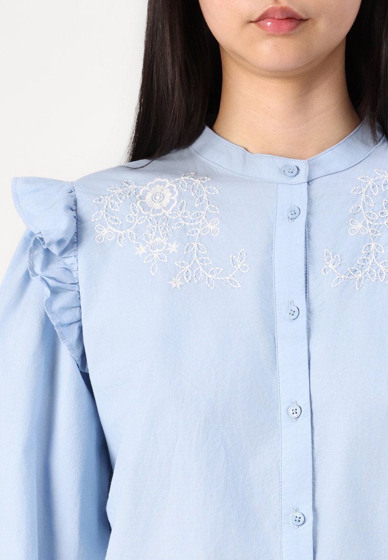 Chemise boutonnée bleu clair avec des détails de volants aux épaules et une broderie florale sur la poitrine. Tissu doux avec une texture lisse.