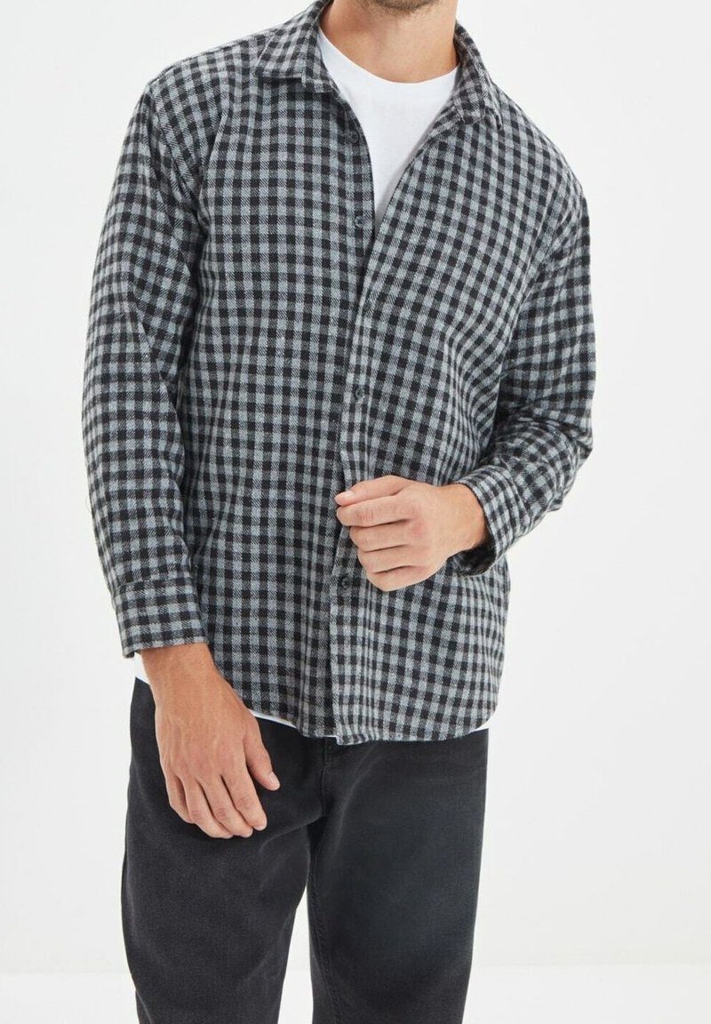Homme portant une chemise à carreaux noire et grise boutonnée par-dessus un t-shirt blanc avec un pantalon noir, debout devant un fond blanc.
