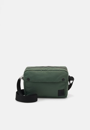 Sac bandoulière en nylon vert avec fermeture éclair, sangle noire réglable, poche avant et un patch logo sur le côté. Texture lisse.