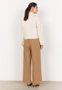 Veste courte beige à manches longues, avec des poignets boutonnés et un col, associée à un pantalon marron ample et des escarpins rayés noir et blanc.