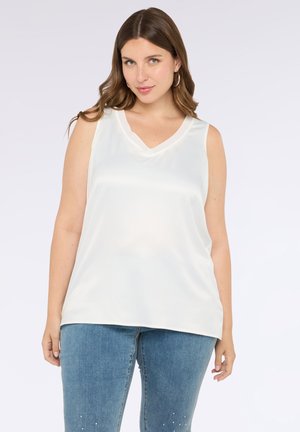 Haut sans manches blanc avec un décolleté en V et une texture lisse et brillante. Assorti à un jean bleu clair avec des accents décoratifs près du bas.