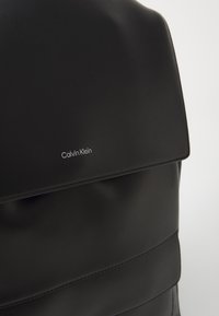 Zaino in pelle nera con texture liscia, patta pieghevole e distintivo marchio Calvin Klein in argento.