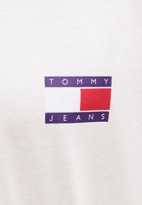Tecido branco de algodão com um logotipo retangular em roxo, vermelho e branco, com o texto "TOMMY JEANS" em letras maiúsculas e a negrito.