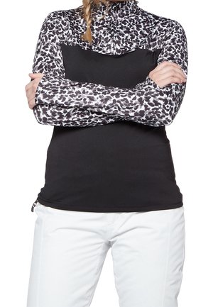 Žena ve černobílém dlouhém tričku s leopardím vzorem a zipem u krku, se zkříženýma rukama a bílými kalhotami.