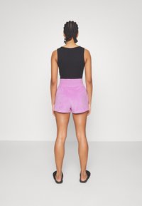 Svart ärmlös topp, rosa velourshorts med hög midja, bärs med svarta slip-in-sandaler, sedd bakifrån mot en vit bakgrund.
