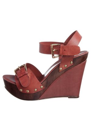 Czerwone skórzane sandały na koturnie z paskiem wokół kostki, złotymi klamrami oraz okrągłymi ćwiekami. Koturn o fakturze drewna.