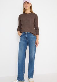 Brauner gerippter Pullover, blaue Schlagjeans und weiße Sneakers. Hellrosa Kappe und lässige Passform. Vertikale Nähte an den Jeans setzen Akzente.