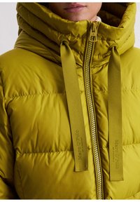 Olijfgroene gestikte jas met een hoge kraag, ritssluiting aan de voorkant en twee getextureerde stoffen trekkoorden met een reliëflogo.