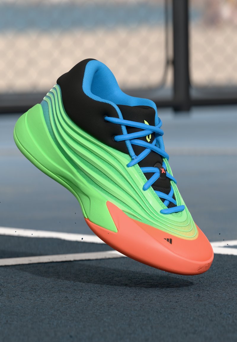 Jasnozielone i pomarańczowe sportowe sneakersy z niebieskimi sznurówkami i czarną kołnierzówką, unoszące się nad niebieską powierzchnią boiska sportowego w pobliżu białych linii.