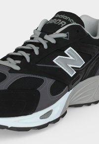 Μαύρο και γκρι αθλητικό παπούτσι New Balance με ανοιχτό γκρι κορδόνια, λευκή και γκρι μεσαία σόλα και μεγάλο λογότυπο "N" στην πλαϊνή πλευρά.