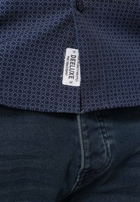 Marineblauwe geblokte overhemd met cirkelvormige ontwerpen, textuur van de stof zichtbaar, etiket leest "DEELUXE 74." Gecombineerd met donkere denim jeans.