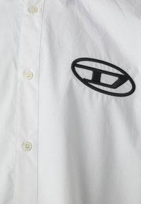 Chemise blanche à manches longues avec un logo noir brodé. Fabriquée en tissu lisse, avec un col classique et une coupe régulière.
