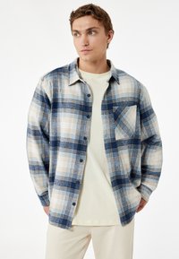 Sininen ja beige ruutukuviollinen flanellipaita, jossa on napitettava etuosa, rintatasku ja rento istuvuus. Kerroksittain vaalean värisen t-paidan päällä.