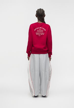 Personne aux cheveux tressés portant un sweat-shirt rouge Nike avec l'inscription "ATHLETICS Nike Department Since 1972" et un pantalon large gris avec un liseré rouge.
