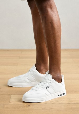 Bele teniske iz sintetičnega materiala z oblazinjenim jezikom, teksturiranimi stranskimi paneli in črnim logotipom na strani. Ravna gumijasta podplat.