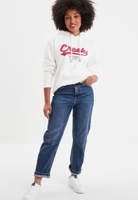 Felpa con cappuccio crema e logo rosso "Creeks", abbinata a jeans di denim blu e sneakers bianche. Il tessuto appare morbido con una vestibilità casual.