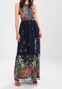 Ärmlös maxiklänning med åtsittande blommig bodice och marinblå kjol med flerfärgade blommönster, bärs med röda öppna klackskor.