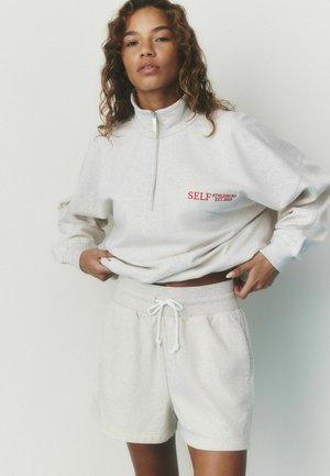 Woman with curly hair wearing light beige zip-up sweatshirt and matching shorts, adjusting sweatshirt with hands.