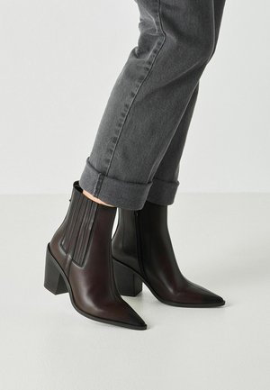 Person trägt dunkelbraune Stiefeletten mit spitzer Spitze, Blockabsatz und schwarzen elastischen Seitenteilen, kombiniert mit hochgekrempelten dunkelgrauen Jeans.