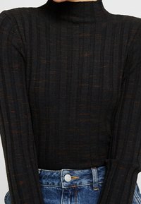 Haut à col roulé côtelé noir avec un col montant, présentant des accents métalliques subtils, associé à un jean en denim taille haute.