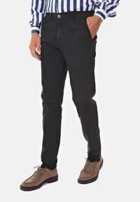 Pantaloni neri slim-fit realizzati in tessuto resistente, con chiusura a bottone frontale e tasche laterali, abbinati a scarpe in pelle scamosciata di colore marrone chiaro.