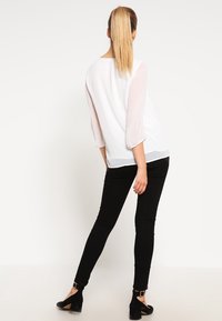 Top bianco con maniche lunghe trasparenti, abbinato a jeans skinny neri e tacchi a cinturino neri. Tessuto morbido, vestibilità casual e design minimale.