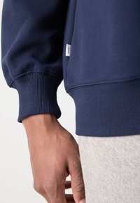 Gros plan sur la main d'une personne à côté d'un sweat-shirt bleu marine à bord-côtes et d'un pantalon gris clair texturé.