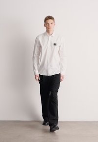 Λευκό πουκάμισο με μακριά μανίκια και κουμπιά, με μικρή μαύρη ετικέτα στο αριστερό στήθος, σε συνδυασμό με μαύρο παντελόνι και μαύρα παπούτσια, σε ουδέτερο περιβάλλον.