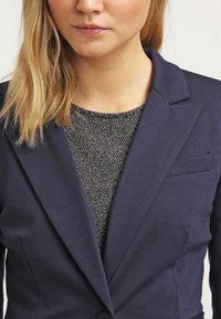 Blazer navy con solapa muesca, cierre de un botón y un discreto detalle de bolsillo frontal, superpuesto a una parte superior oscura texturizada.
