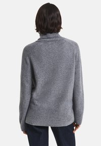 Persona con capelli scuri di lunghezza spalle che indossa un maglione grigio a collo alto largo e pantaloni scuri, vista da dietro.