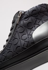 Černé textilní tenisky s embosovaným vzorem loga, zipovým zapínáním a lesklým černým akcentem na patě. Texturovaný povrch dodává hloubku.