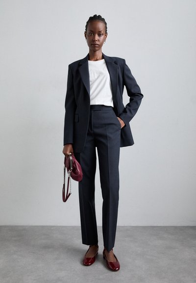 Costum tailored bleumarin cu sacou structurat și pantaloni slim-fit, asociat cu un top alb și o poșetă burgundă. Pantofii roșii lucioși completează ținuta.
