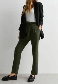 Pantaloni verdi a vita alta con una vestibilità sartoriale, tasche laterali e cintura in tessuto, abbinati a una maglietta bianca aderente e a un blazer nero. Mocassini neri.
