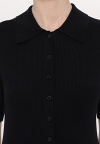 Czarny kardigan z prążkowanej dzianiny z krótkimi rękawami, okrągłym kołnierzem i sześcioma pasującymi guzikami z przodu. Miękka faktura i dopasowany kształt.