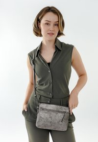 Borsa a tracolla grigio metallico con superficie texturizzata, dotata di tasca frontale con zip e tracolla regolabile, abbinata a un outfit verde oliva.